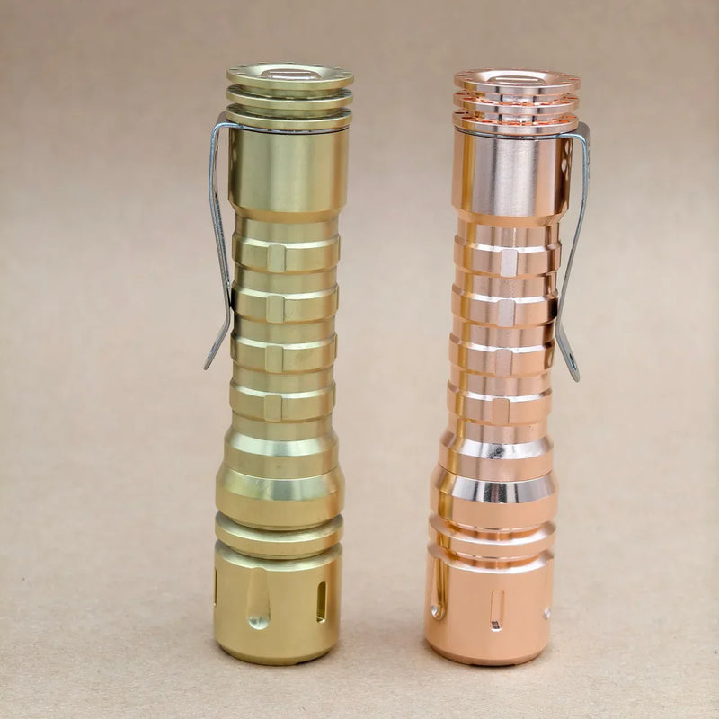 Two ReyLight LANapple flashlights with ribbed bodies and pocket clips—a brass (gold-colored) and a copper-colored—stand upright on a beige surface. Both feature high CRI LED illumination.