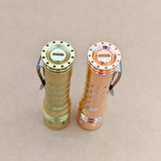 A ReyLight LANapple - Brass flashlight and a copper model stand upright side by side on a beige surface, seen from above. Both deliver high CRI LED illumination.