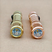 Two metallic flashlights, including the ReyLight LANapple - Copper with a Nichia 519a 4000K R9080 LED, are placed side by side on a brown surface with their lenses facing forward.