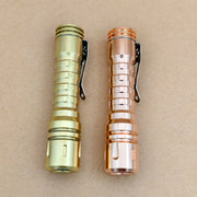 Two metal flashlights, a ReyLight LANapple - Copper with Nichia 519a 4000K R9080 LED and a gold one, both with black clips, are placed side by side on a plain brown surface.