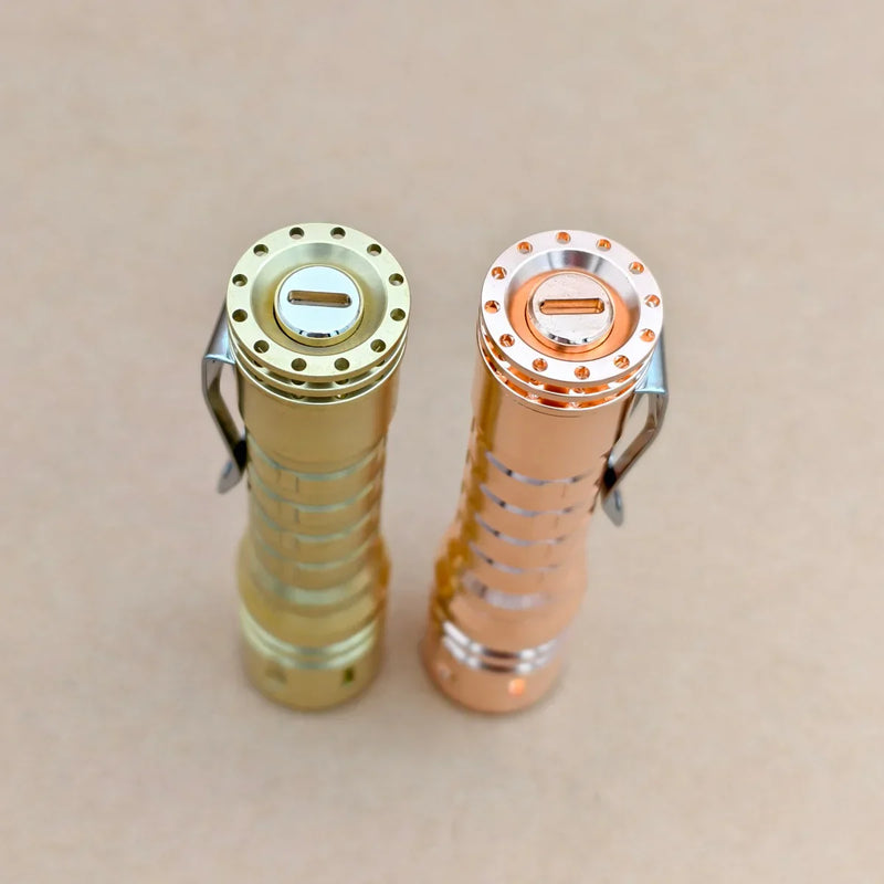 Two metal flashlights, including the ReyLight LANapple - Copper with a Nichia 519a 4000K R9080 LED, stand upright side by side on a beige surface, viewed from above.
