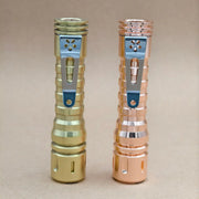 Two metallic flashlights, including a ReyLight LANapple - Copper with a Nichia 519a 4000K R9080 LED, stand upright on a plain beige surface against a neutral background.