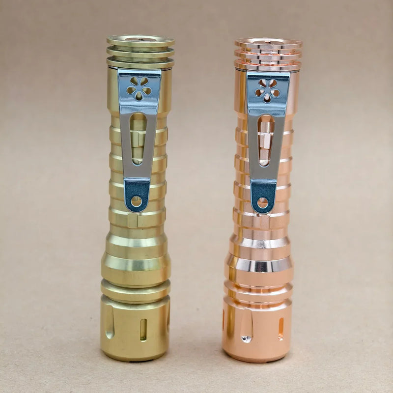 Two metallic flashlights, including a ReyLight LANapple - Copper with a Nichia 519a 4000K R9080 LED, stand upright on a plain beige surface against a neutral background.