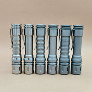 Six metallic flashlights with pocket clips, including the ReyLight LANapple - Ti, are standing upright in a row against a plain beige background. Some models support rechargeable 14500 batteries.