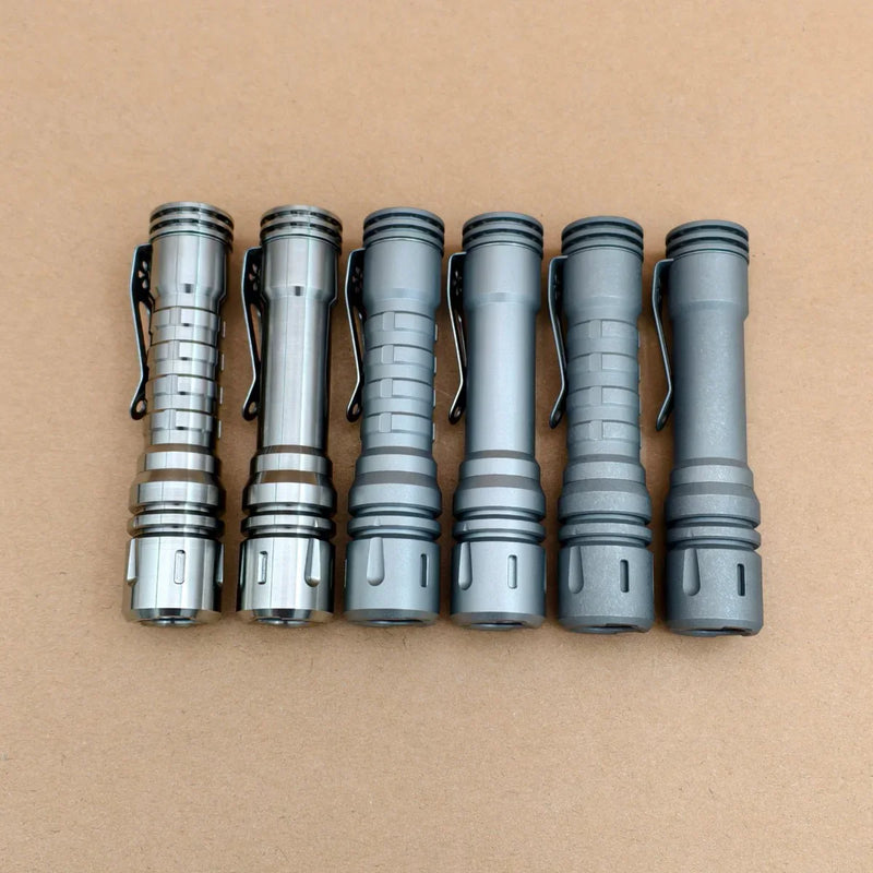 Six grey and metallic flashlights, including the ReyLight LANapple - Ti, are lined up side by side on a plain brown surface, each compatible with a rechargeable 14500 battery.