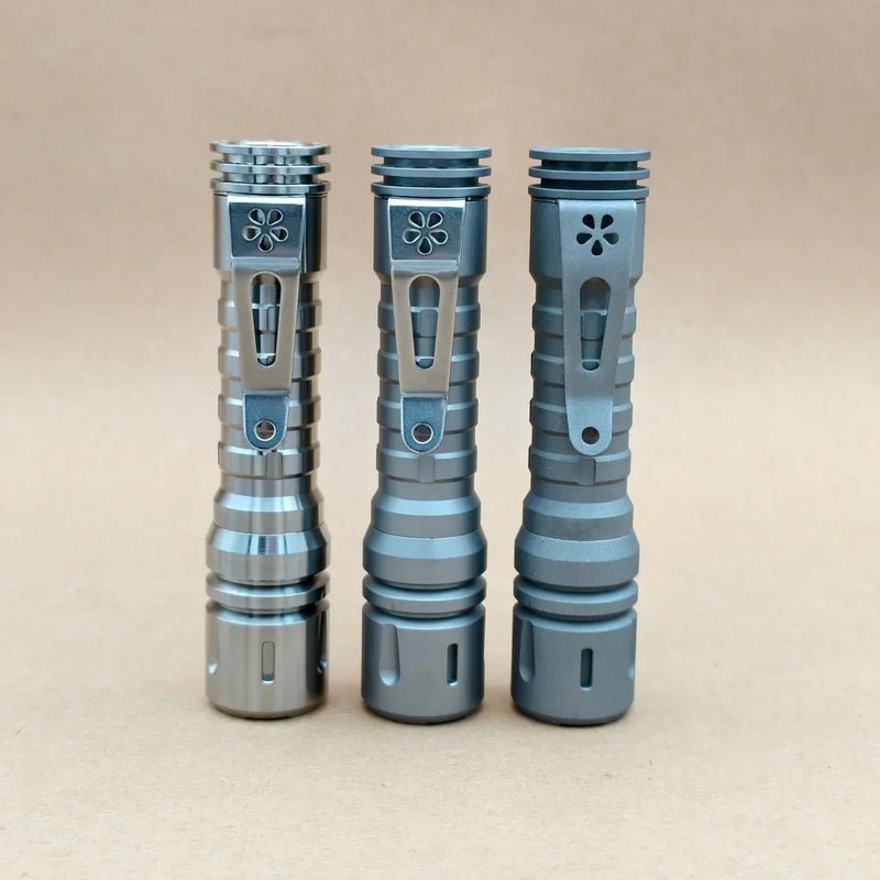 Three ReyLight LANapple - Ti flashlights with grooved titanium bodies and flower-shaped clips stand upright on a light brown surface, each compatible with a rechargeable 14500 battery, against a plain background.