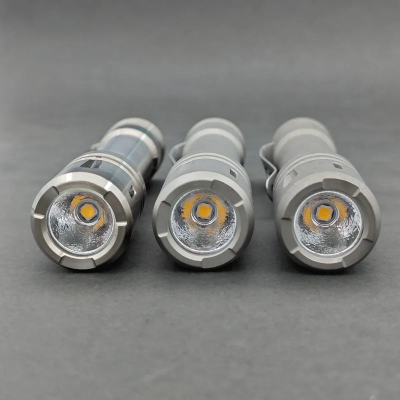 Three ReyLight LANapple - Ti flashlights in gray are side by side on a gray surface, displaying their LED bulbs and reflectors. Each flashlight operates with a rechargeable 14500 battery for dependable lighting.