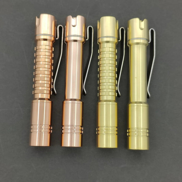 Four tactical pens in bronze and gold colors with different textures and pocket clips, displayed on a dark surface alongside a ReyLight Pineapple Mini Brass Flashlight.