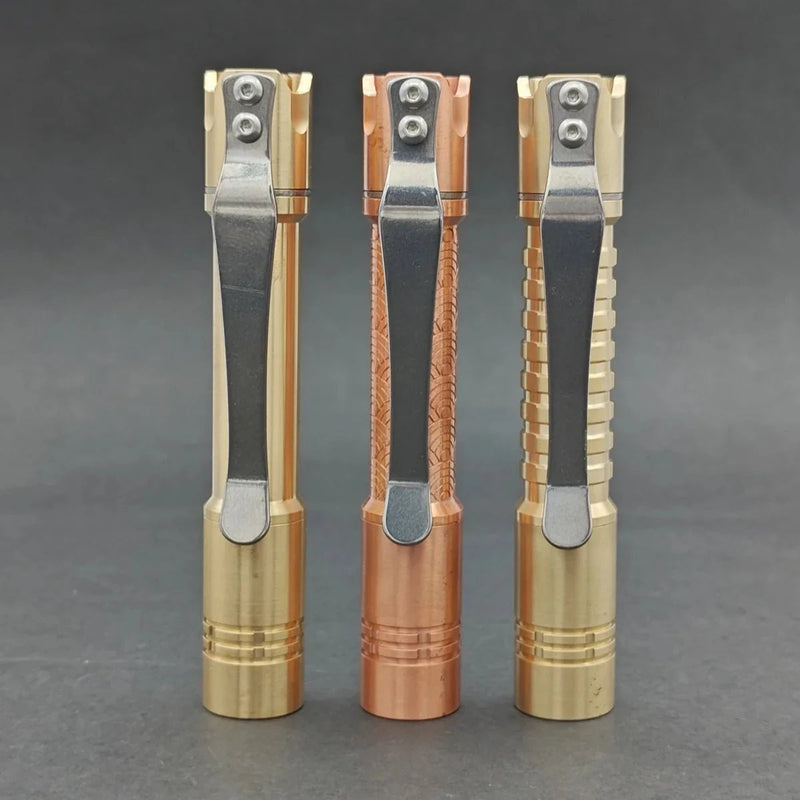 Three metallic flashlights, including a ReyLight Pineapple Mini MK II - Copper&Brass with metal pocket clips, stand upright side by side against a dark background.