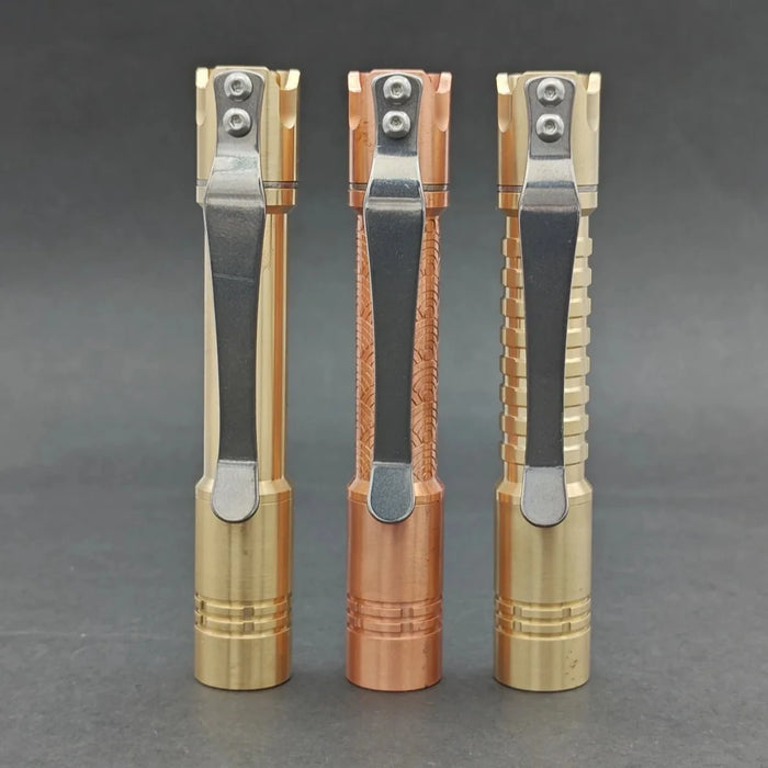 Three ReyLight Pineapple Mini MK II flashlights, with copper and brass textured bodies and silver clips, stand upright on a gray surface. Each uses the Nichia 519a LED for exceptional brightness. Compatible with a rechargeable 10440 battery, they effortlessly illuminate any setting.