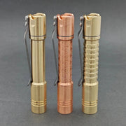 Three metal flashlights, including the ReyLight Pineapple Mini MK II in copper and brass with a Nichia 519a LED, stand upright in gold and copper tones on a gray background, each featuring distinct surface patterns.