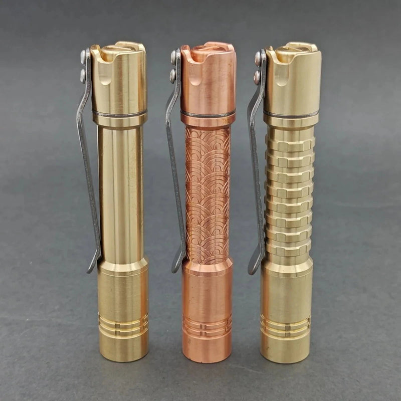 Three metal flashlights, including the ReyLight Pineapple Mini MK II in copper and brass with a Nichia 519a LED, stand upright in gold and copper tones on a gray background, each featuring distinct surface patterns.