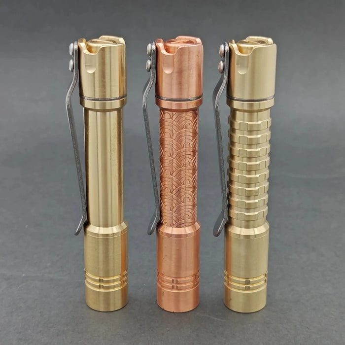 Three metal flashlights, including the stylish ReyLight Pineapple Mini MK II in copper and brass, stand upright on a gray surface. Their distinct textures range from smooth to engraved and ribbed, with gold-tone and copper-tone finishes. Each features a rechargeable 10440 battery and Nichia 519a LED for top performance.