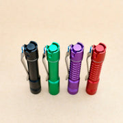 Four ReyLight Pineapple Mini MK3-Aluminum flashlights in black, green, purple, and red stand upright with pocket clips on a plain beige background. Each uses a rechargeable 10440 battery for portable convenience.