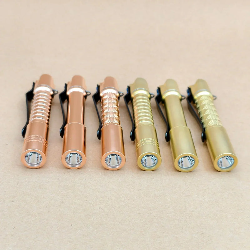 Six compact ReyLight Pineapple Mini MK 3 flashlights in copper and brass are arranged in a row on a neutral surface, all facing forward.