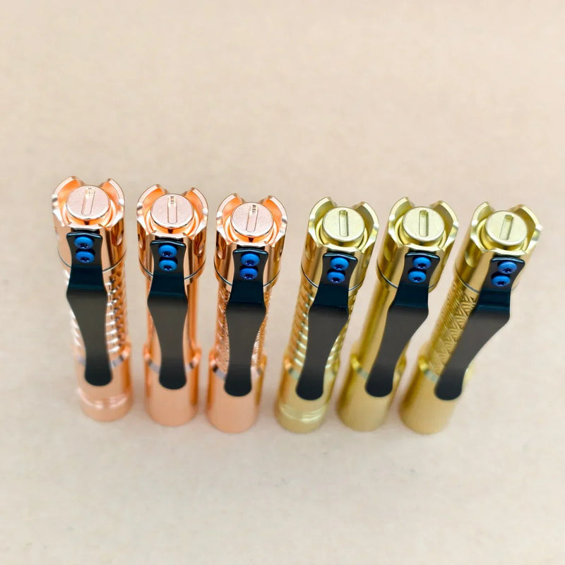 Three copper and three gold ReyLight Pineapple Mini MK 3 flashlights, including a brass model, are standing upright in a row on a beige surface, viewed from above.
