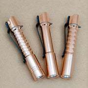 Three ReyLight Pineapple Mini MK 3 - Copper&Brass flashlights with black clips are arranged side by side on a brown surface; each features a unique body pattern.