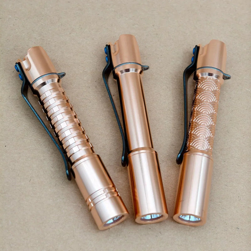 Three ReyLight Pineapple Mini MK 3 - Copper&Brass flashlights with black clips are arranged side by side on a brown surface; each features a unique body pattern.