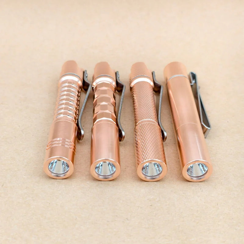 Four ReyLight Pineapple Mini MK3 flashlights in copper and brass, each featuring distinct grip textures, are arranged side by side on a plain tan surface, highlighting the sleek appeal and practical design that define high-quality copper EDC flashlights.