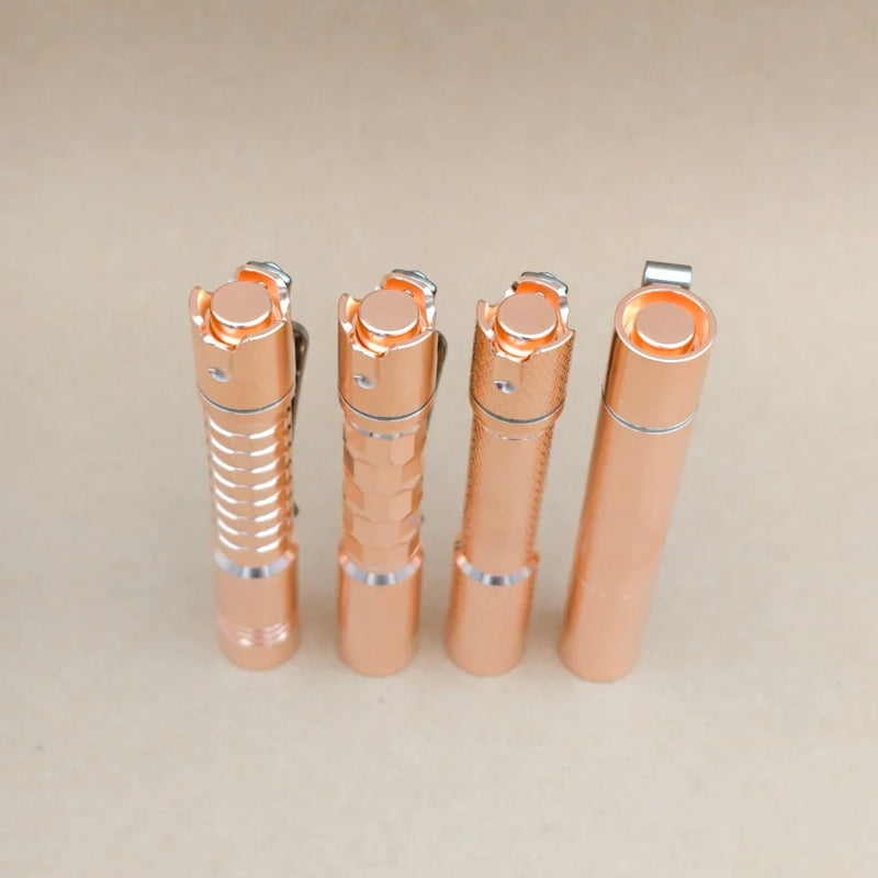 Four ReyLight Pineapple Mini MK3 flashlights in copper and brass finishes, each featuring a unique textured grip, are standing upright in a row on a light beige surface.