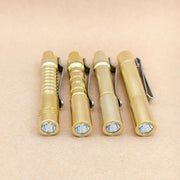 Four ReyLight Pineapple Mini MK3 flashlights—crafted from copper and brass with distinct textured grips—are lined up side by side on a light brown surface, each featuring a pocket clip and their lenses facing forward. Designed for everyday carry, these EDC lights blend style and functionality, making them an appealing choice for enthusiasts who value quality metals like copper and brass in their gear.