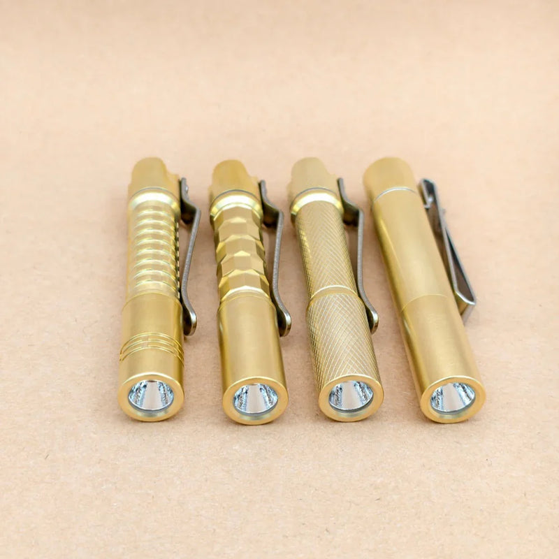 Four ReyLight Pineapple Mini MK3 flashlights—crafted from copper and brass with distinct textured grips—are lined up side by side on a light brown surface, each featuring a pocket clip and their lenses facing forward. Designed for everyday carry, these EDC lights blend style and functionality, making them an appealing choice for enthusiasts who value quality metals like copper and brass in their gear.