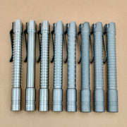 Seven metallic pen-shaped flashlights with pocket clips, including the rechargeable ReyLight Pineapple Mini Penlight - TI, are lined up on a plain brown surface, each displaying a distinct textured grip pattern.