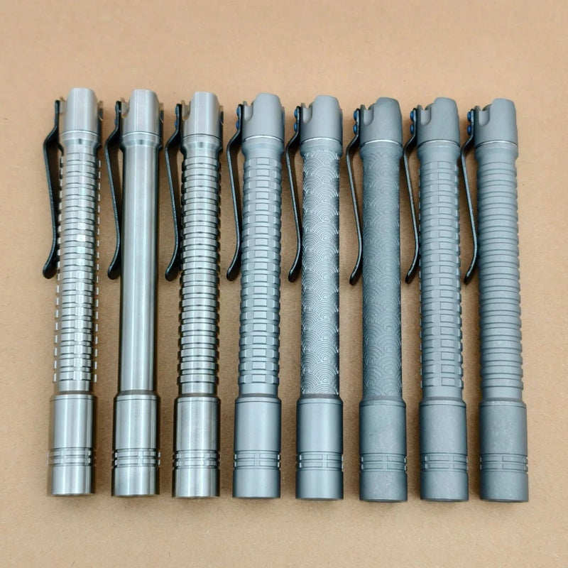 Seven metallic pen-shaped flashlights with pocket clips, including the rechargeable ReyLight Pineapple Mini Penlight - TI, are lined up on a plain brown surface, each displaying a distinct textured grip pattern.