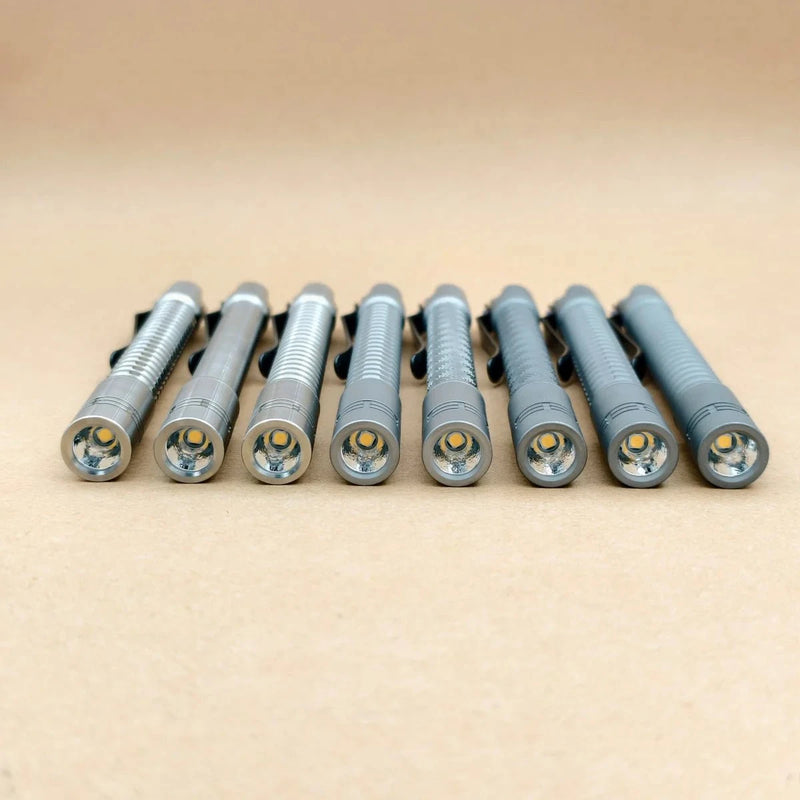 Eight metallic flashlights, including the ReyLight Pineapple Mini Penlight - TI, are lined up in a row on a beige surface with their lenses facing forward.