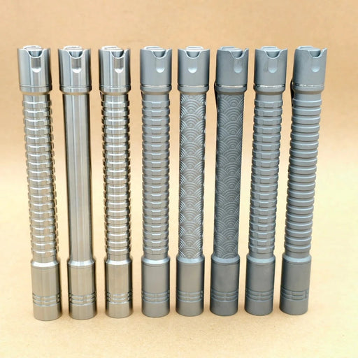 Seven metallic cylindrical penlights, including the ReyLight Pineapple Mini Penlight - TI with textured grips, stand upright in a row against a plain light brown background. One resembles a rechargeable 500-lumen flashlight or penlight.