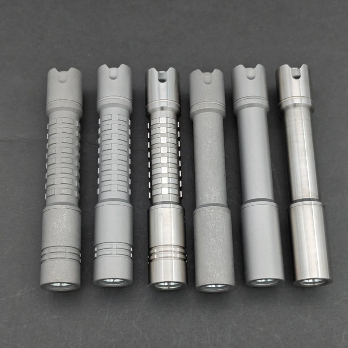 Five metallic flashlights, including the sleek ReyLight Pineapple Mini Ti - MK II with distinct grip patterns, are lined up vertically on a dark gray surface. Featuring Nichia 519a R9080 LEDs and powered by USB-C 10440 batteries, they ensure both style and performance.