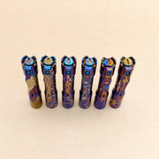 Six ReyLight Pineapple Mini Ti MK3 flashlights, each hand carved and heat anodized with unique patterns, stand upright in a row on a plain beige background.