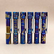 Six metallic pens, including the ReyLight Pineapple Mini Ti MK3—hand carved and heat anodized with gold and blue patterns—stand upright in a row against a plain, light brown background.