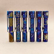Six hand-carved, heat-anodized ReyLight Pineapple Mini Ti MK3 pens with detailed blue and gold patterns stand upright in a row against a plain beige background.
