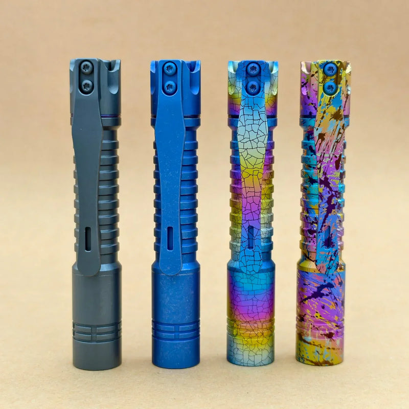 Four ReyLight Pineapple Mini Titanium Frag - Anodized flashlights stand upright in a row on a beige surface; two feature solid blue tones, while the other two display vibrant, patterned finishes.