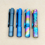 Four ReyLight Pineapple Mini Titanium Frag - Anodized flashlights in matte grey, matte blue, rainbow gradient, and a multicolored cracked finish are arranged vertically on a plain beige surface.
