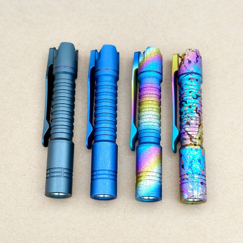 Four ReyLight Pineapple Mini Titanium Frag - Anodized flashlights in matte grey, matte blue, rainbow gradient, and a multicolored cracked finish are arranged vertically on a plain beige surface.
