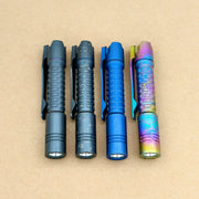 Four ReyLight Pineapple Mini Titanium Frag - Anodized flashlights, each with a different color and finish, featuring textured grips and clips, are arranged side by side on a plain brown background.