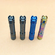 Four ReyLight Pineapple Mini Titanium Frag - Anodized flashlights, each with a different finish—black, speckled gray, blue, and multicolored—stand upright on a light brown surface.