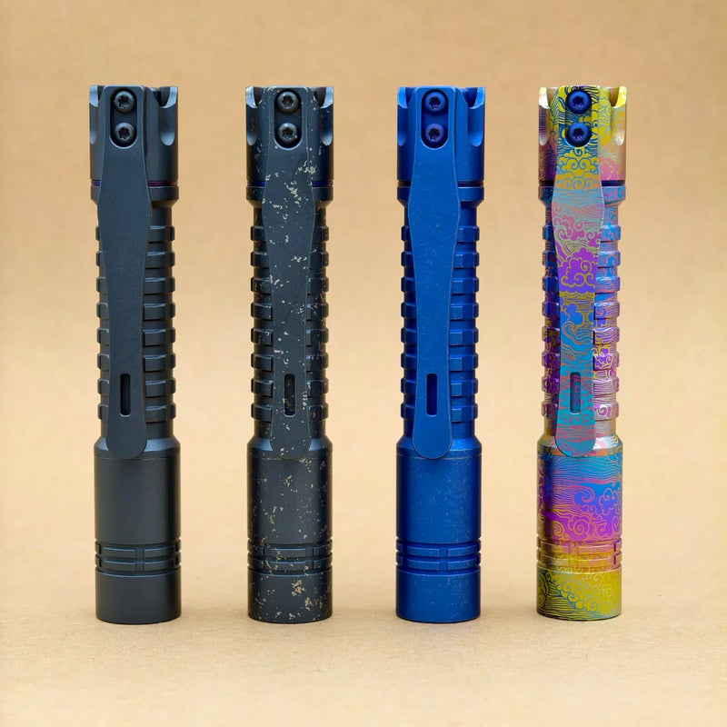 Four ReyLight Pineapple Mini Titanium Frag - Anodized flashlights are standing upright in a row on a plain tan background, each with a unique finish: black, speckled black, blue, and multicolored with patterns.