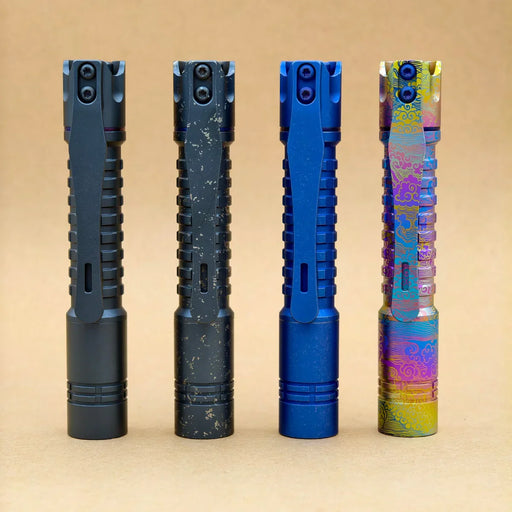Four ReyLight Pineapple Mini Titanium Frag - Anodized flashlights are standing upright in a row on a plain tan background, each with a unique finish: black, speckled black, blue, and multicolored with patterns.