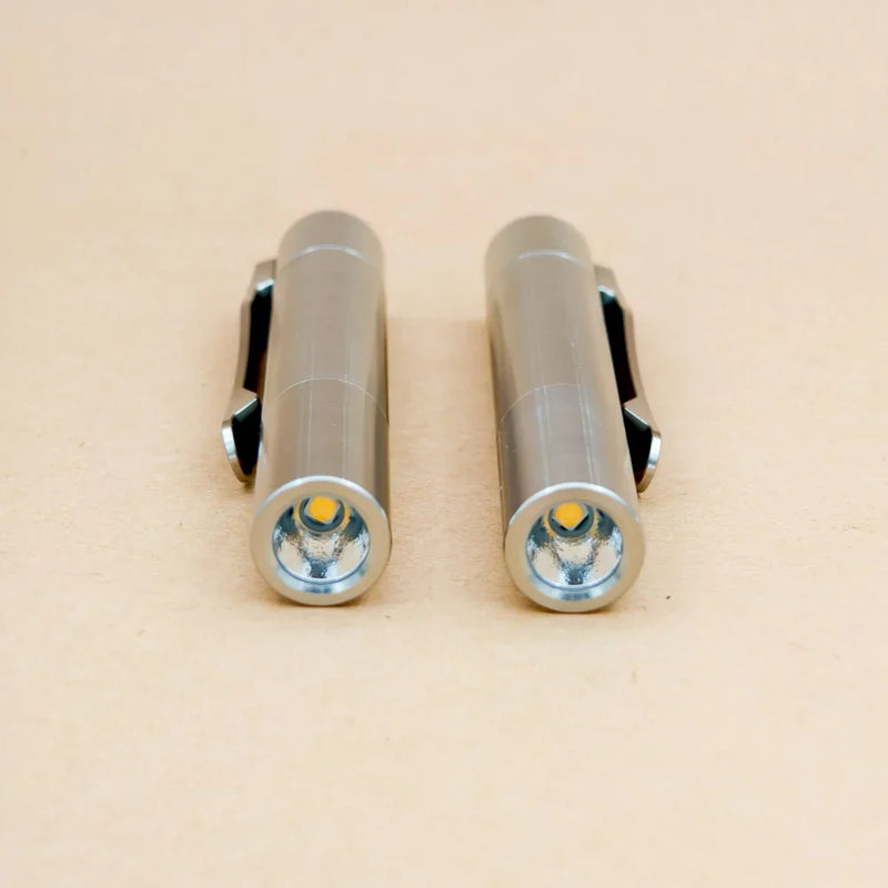 Two ReyLight Pineapple Mini Titanium MK3 - Cylinder flashlights with clips, finished in small silver and featuring Ti raw screws, are positioned side by side on a light beige surface, both facing forward.