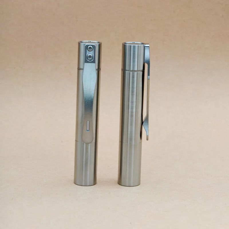 Two ReyLight Pineapple Mini Titanium MK3 - Cylinder pens stand upright on a beige surface; one reveals its clip, while the other highlights a button and small window, showcasing their sleek cylindrical design.