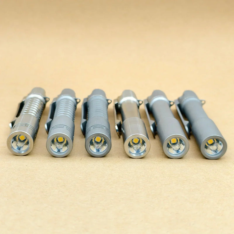 Six ReyLight Pineapple Mini Titanium MK3 flashlights—compact and ideal for everyday carry—are lined up side by side on a plain beige surface, showcasing their lenses and high-CRI LED bulbs.