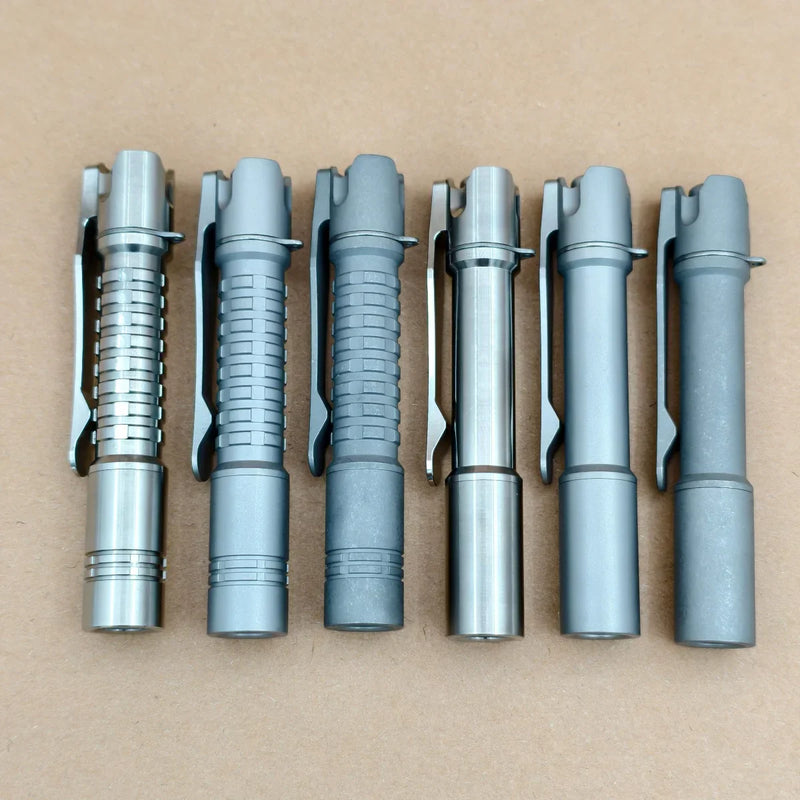 Six ReyLight Pineapple Mini Titanium - MK3 pen-style flashlights, perfect for everyday carry, are lined up on a brown surface; three have grooved grips and three have smooth bodies, all featuring convenient pocket clips.