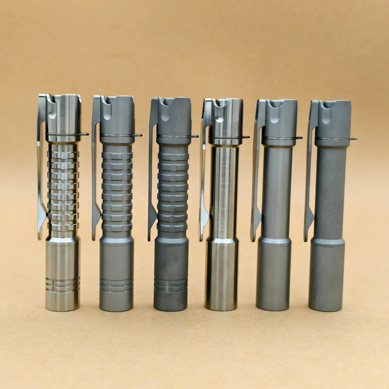 Six ReyLight Pineapple Mini Titanium MK3 pen-style flashlights with pocket clips, including a sleek titanium model, stand upright in a row on a plain tan background—ideal for everyday carry.