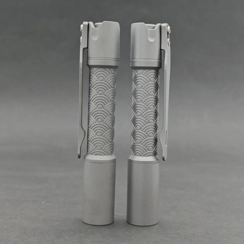 Two ReyLight Pineapple Mini Titanium - MK3 cylindrical flashlights with wave-patterned grips stand upright on a gray surface—stylish and reliable EDC options for any occasion.