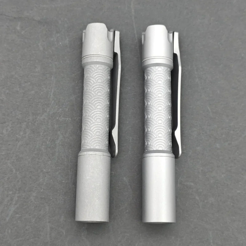 Two identical ReyLight Pineapple Mini Titanium - MK3 pens, featuring textured wave patterns on their barrels, rest side by side on a gray surface, reflecting the sleek design of premium titanium EDC flashlights.