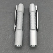 Two ReyLight Pineapple Mini Titanium - MK3 EDC flashlights, featuring textured grips and clips, are placed side by side on a gray surface.