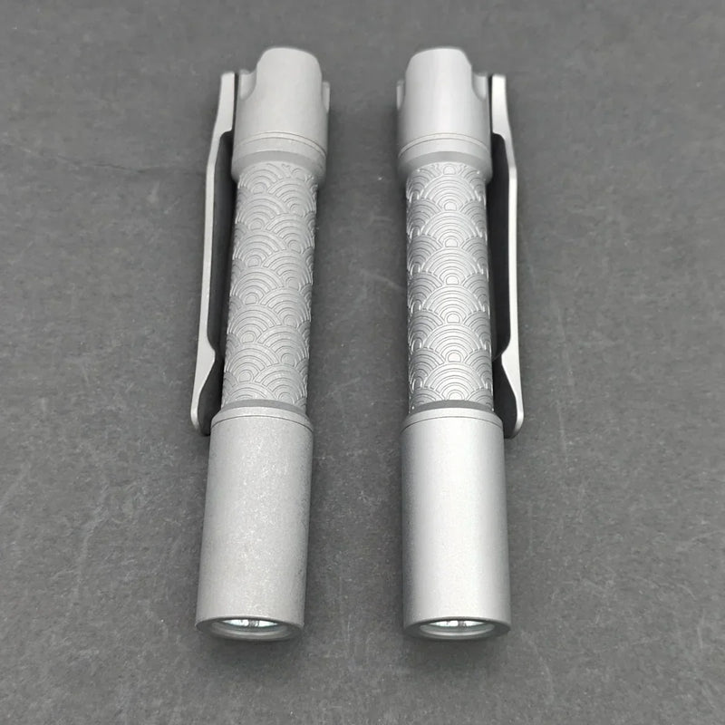 Two ReyLight Pineapple Mini Titanium - MK3 EDC flashlights, featuring textured grips and clips, are placed side by side on a gray surface.